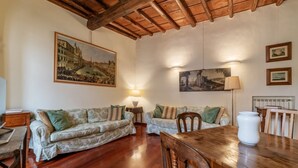 Ein gemütliches Wohnzimmer mit Holzboden, zwei gemusterte Sofas, einem Holzesstisch und einem großen Gemälde eines historischen Gebäudes an der Wand.
