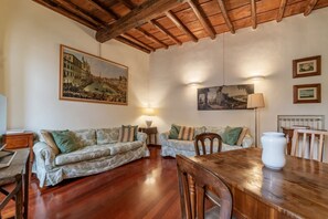A cozy living room with wooden flooring, two patterned sofas, a wooden dining table, and a large painting of a historical building on the wall.