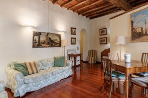 A cozy living room with a wooden ceiling, a sofa with patterned cushions, a dining table with chairs, and framed pictures on the walls.