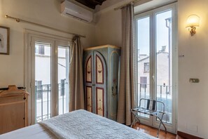 A bedroom with a bed, a wardrobe, a chair, and a balcony with a view of buildings.