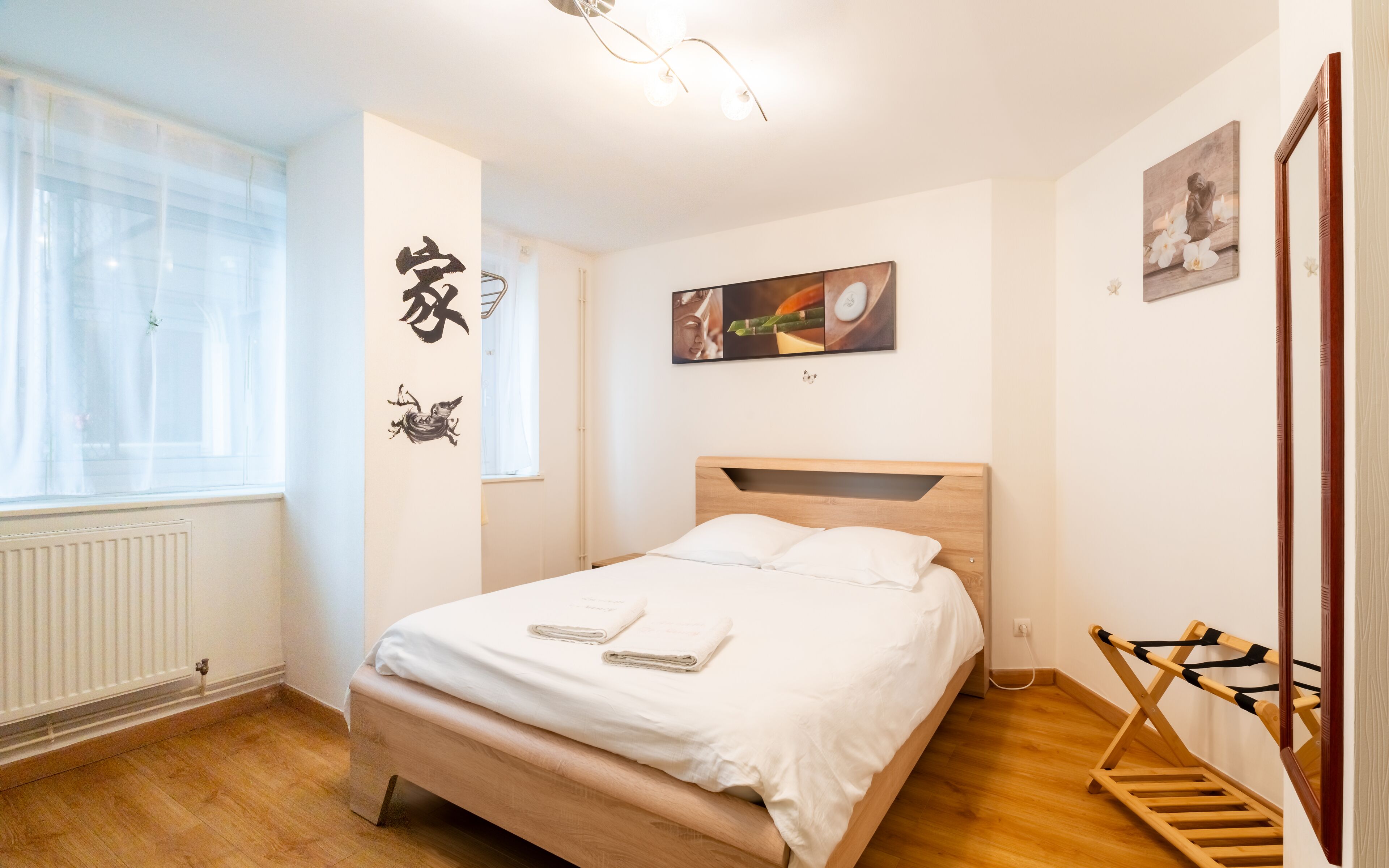 A neatly arranged bedroom with a wooden bed, a radiator, a window with sheer curtains, and a wall-mounted artwork.