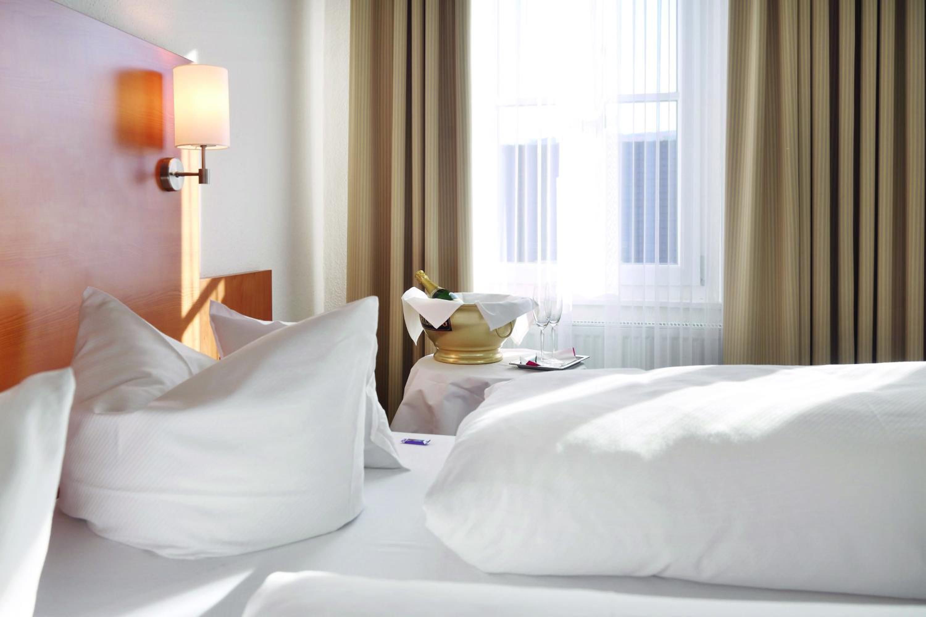 Une chambre d’hôtel avec un lit, des oreillers, une table de chevet, une lampe et une fenêtre avec des rideaux.