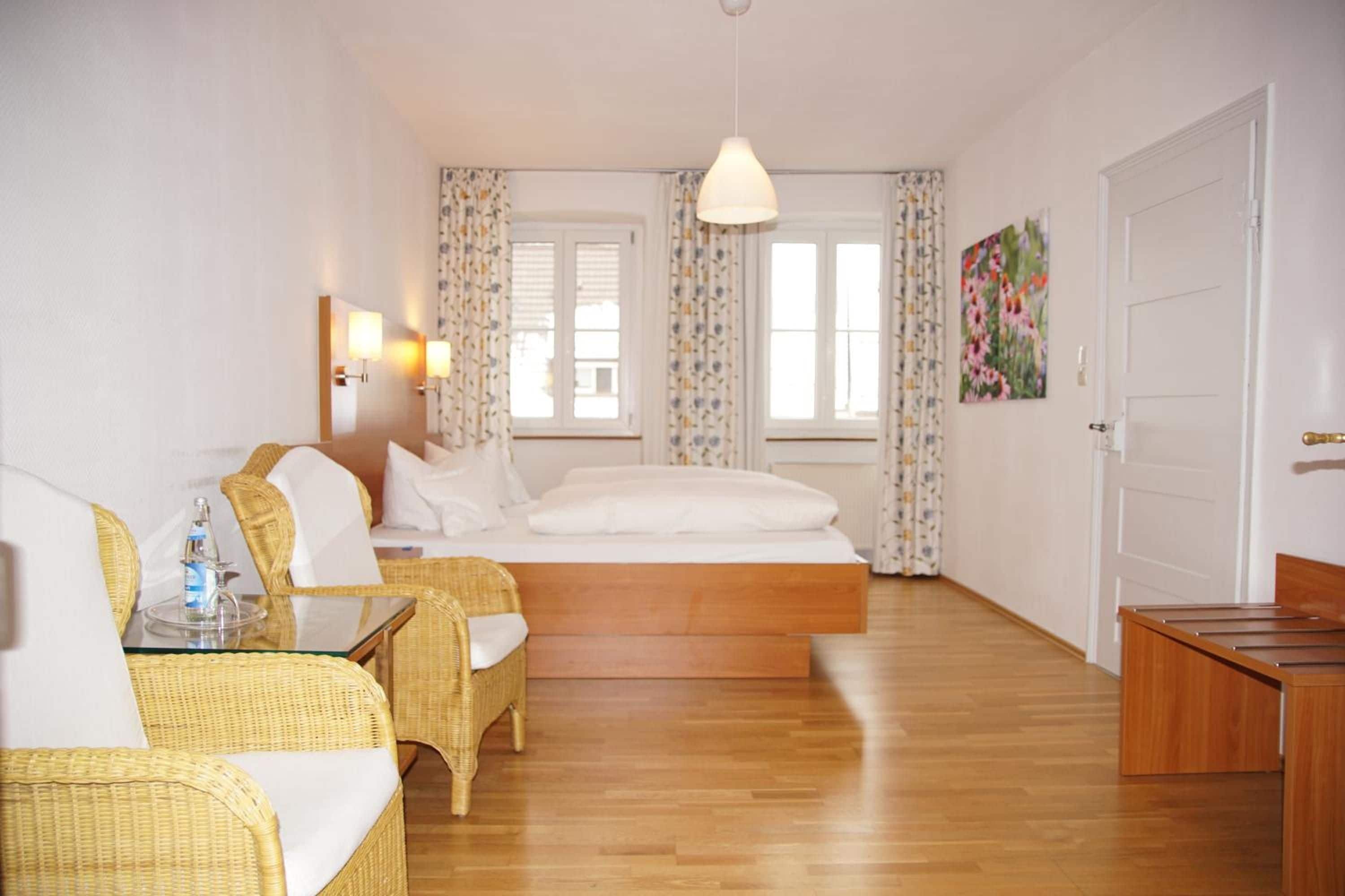 A hotel room with a bed, two wicker chairs, a table with a bottle, and a painting on the wall.