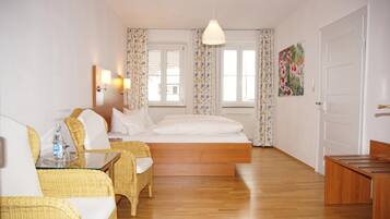 A hotel room with a bed, two wicker chairs, a table with a bottle, and a painting on the wall.