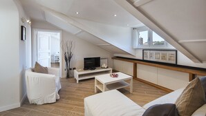 A modern loft apartment with a sloped ceiling, a flat-screen TV on a white console table, a white sofa, and a dining table.