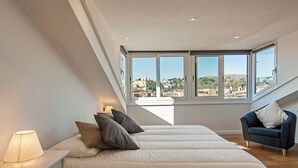 A bedroom with a large window, a bed with pillows, a chair, and a bedside table with a lamp.