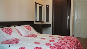 Uma cama bem arrumada com colcha branca e estampa de rosas vermelhas, um espelho sobre a cama e um guarda-roupa de madeira escura.