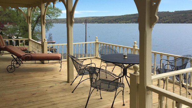 A covered deck with a view of a lake, featuring a table and chairs, and a lounge area with a chair and a small table. - Waterfront Keuka Lake INCREDIBLE VIEWS • BOAT SLIP • TWO BALCONIES 3rd floor (Hammondsport)