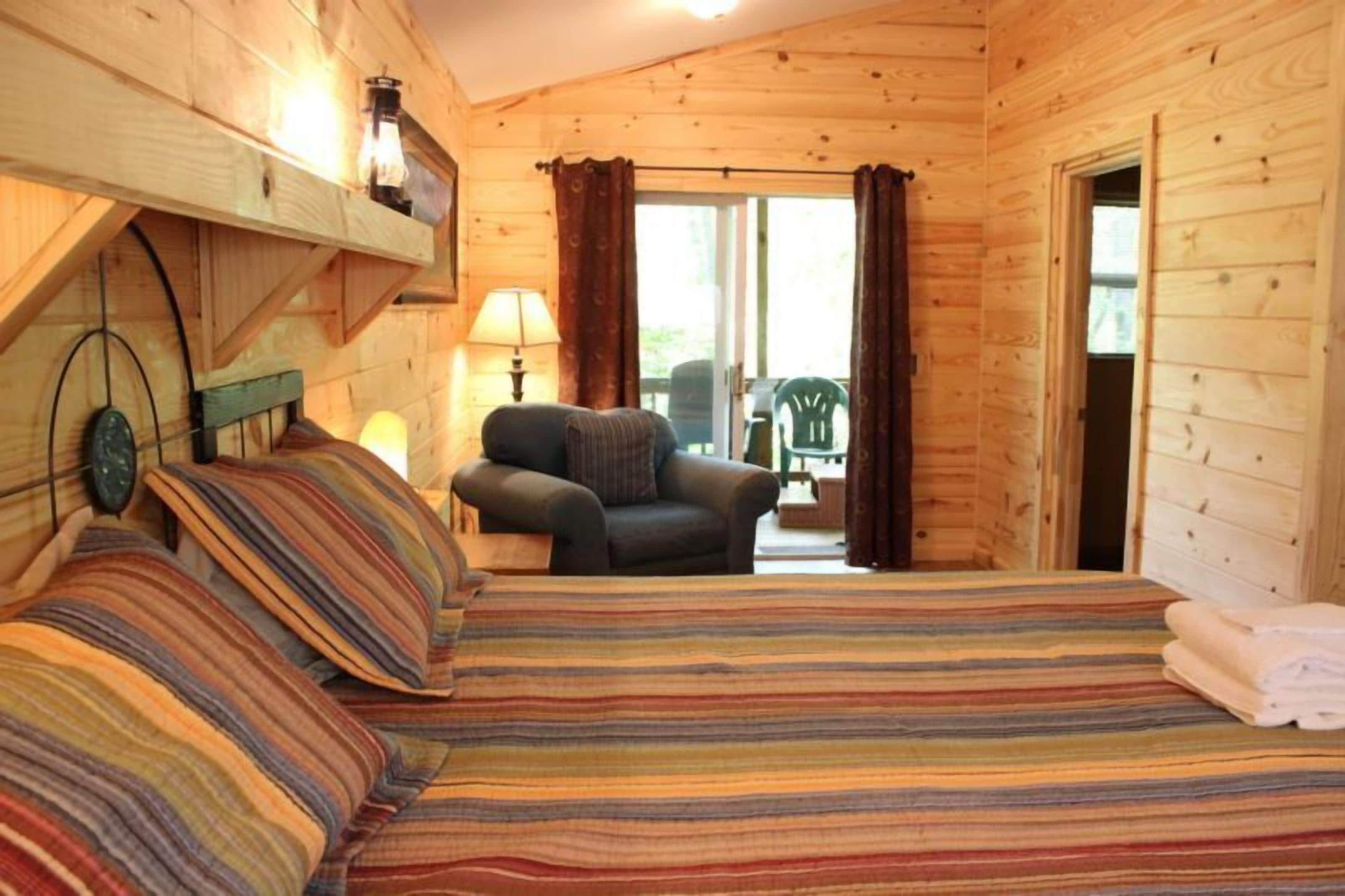A cozy wooden cabin room with a bed, a chair, a lamp, and a view of the outdoors.