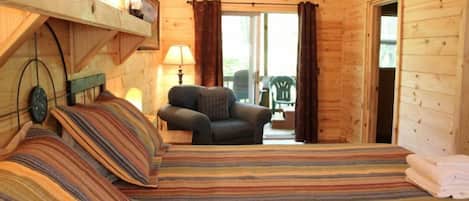 A cozy wooden cabin room with a bed, a chair, a lamp, and a view of the outdoors.
