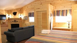 Une pièce chaleureuse aménagée dans un chalet en rondins, avec un canapé, un lit recouvert d’une couverture à rayures, une cheminée et un téléviseur.