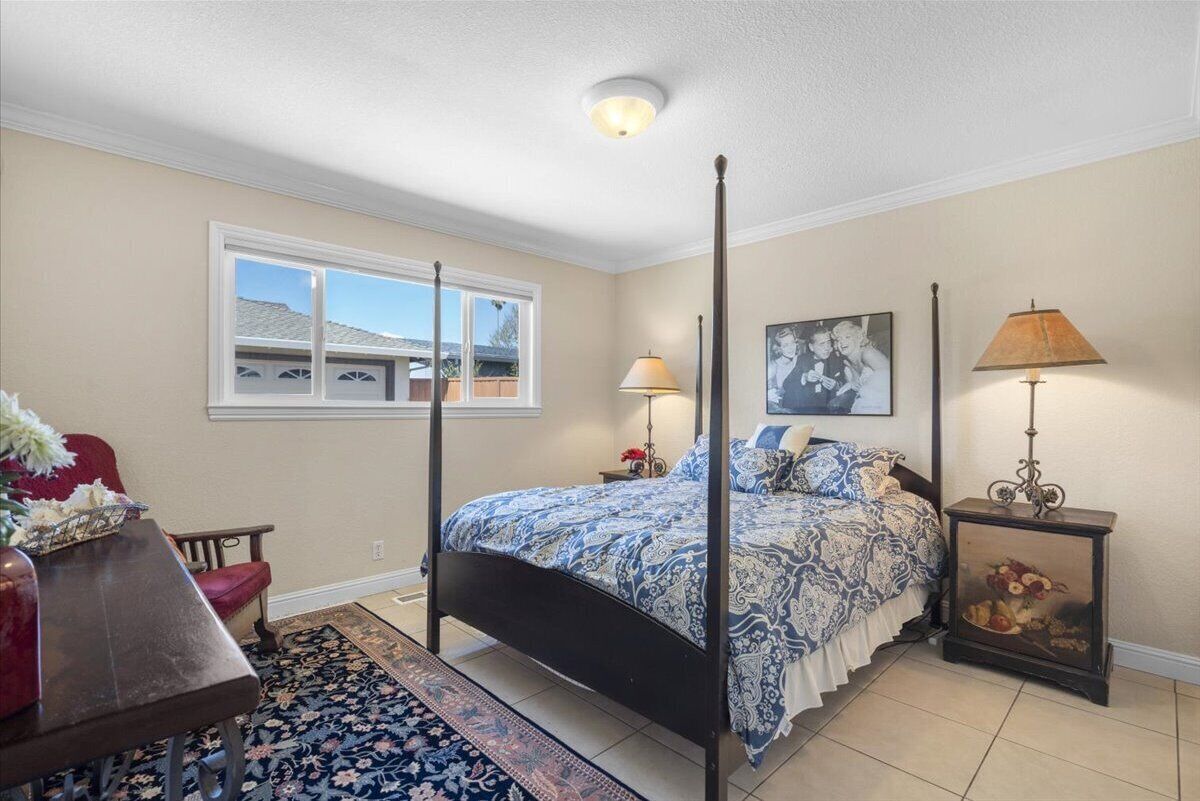 A bedroom with a four-poster bed, a nightstand with a lamp, a chair, a window with a view of a house, and a painting on the wall.