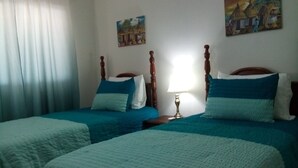 Ein Zimmer mit zwei Betten, beide mit Holz-Kopfteilen und blauer Bettwäsche. Dazwischen steht ein Nachttisch mit einer Lampe.