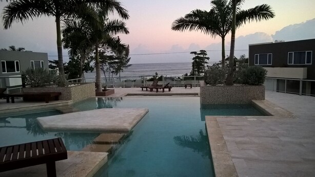 Ein Infinity-Pool mit Meerblick, umgeben von Palmen und modernen Gebäuden.