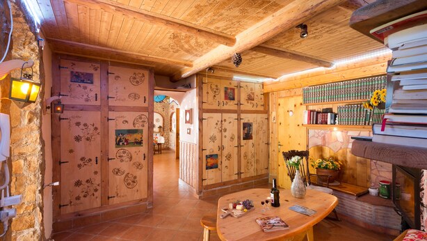Un interior acogedor y rústico con paredes y techos de madera, suelo de baldosas, una mesa de madera con una botella y copas, y una estantería llena de libros y objetos decorativos.