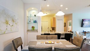 A dining area with a table set for four, chairs, a chandelier, and a kitchen in the background. - Refurbished.  Accessible. Step free access, disabled parking, walk in shower. (Kissimmee)