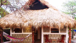 Una cabaña con techo de paja y fachada de ladrillo, dos sillas de madera y una hamaca.