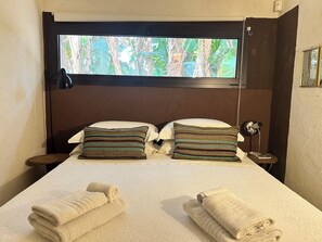 Ein Hotelzimmer mit einem Bett, zwei Kissen, einem Nachttisch, einer Lampe und einem Fenster mit Blick auf Grünflächen.