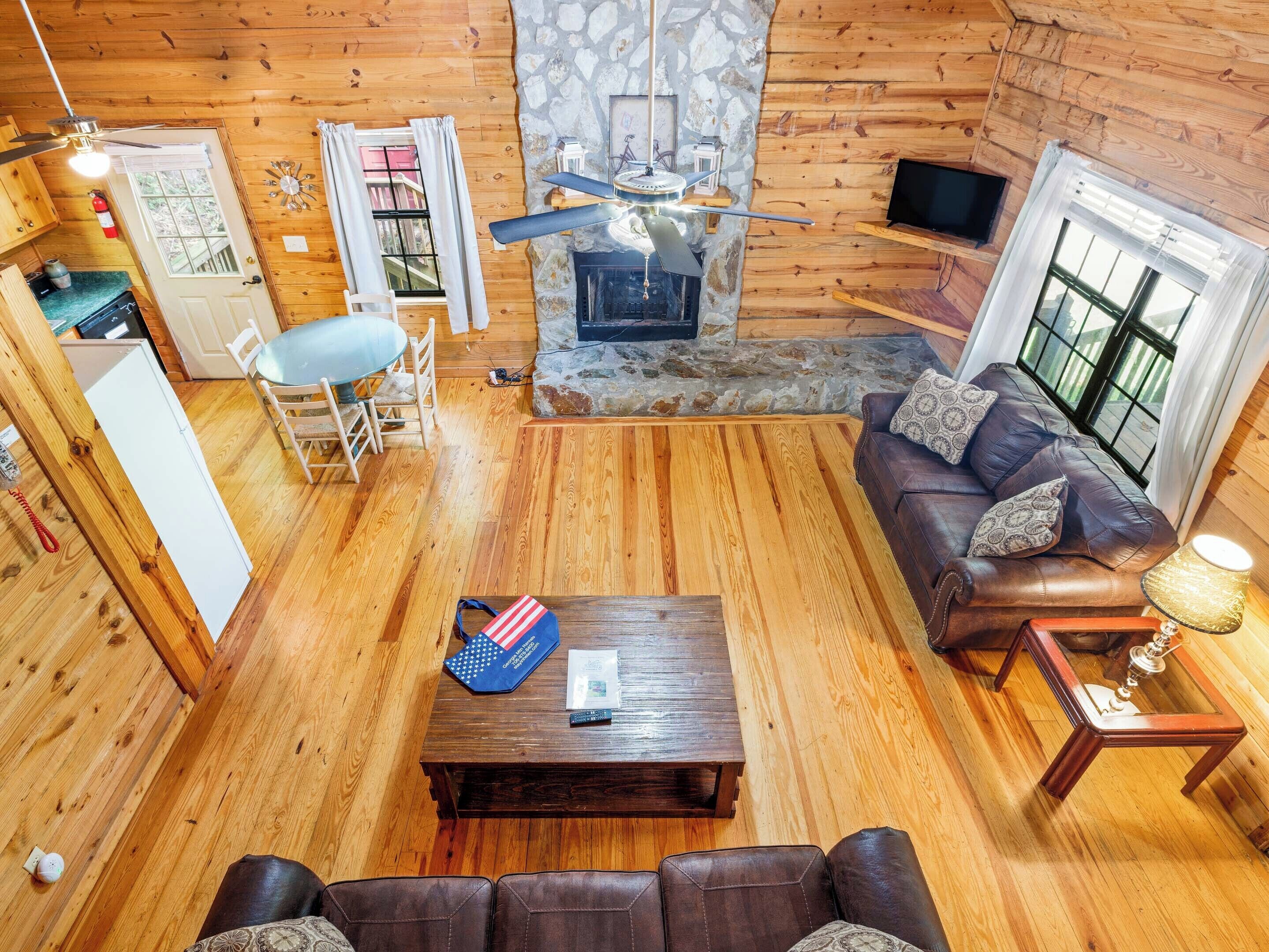 A cozy cabin interior with a stone fireplace, wooden floors, a leather sofa, and a dining area.