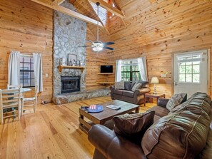 A cozy cabin interior with a stone fireplace, wooden walls, a leather sofa, and a television.