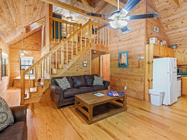 A wooden cabin interior with a staircase, a living room with a leather sofa, a wooden coffee table, and a kitchen area.