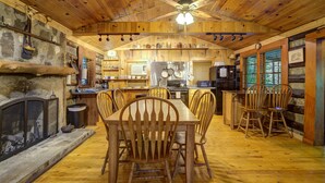A rustic wooden interior with a dining table, chairs, a fireplace, kitchen area, and a ceiling fan. - Adanaylah - A True Log Cabin Experience. Pet Friendly, too! (Hiawassee)