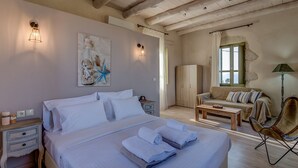 Un dormitorio con una cama grande, mesitas de noche, un banco de madera, una silla y una ventana con cortinas.