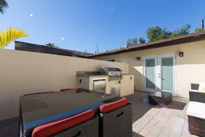 Un patio exterior con una parrilla, una mesa con cojines rojos, sillas de mimbre y un ventanal de vidrio.