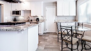 Una cocina moderna con armarios blancos, encimera de granito y comedor con mesa de vidrio y sillas de metal.