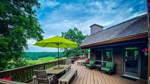 Una terraza de madera con mobiliario exterior, un paraguas amarillo y vistas a un paisaje verde.