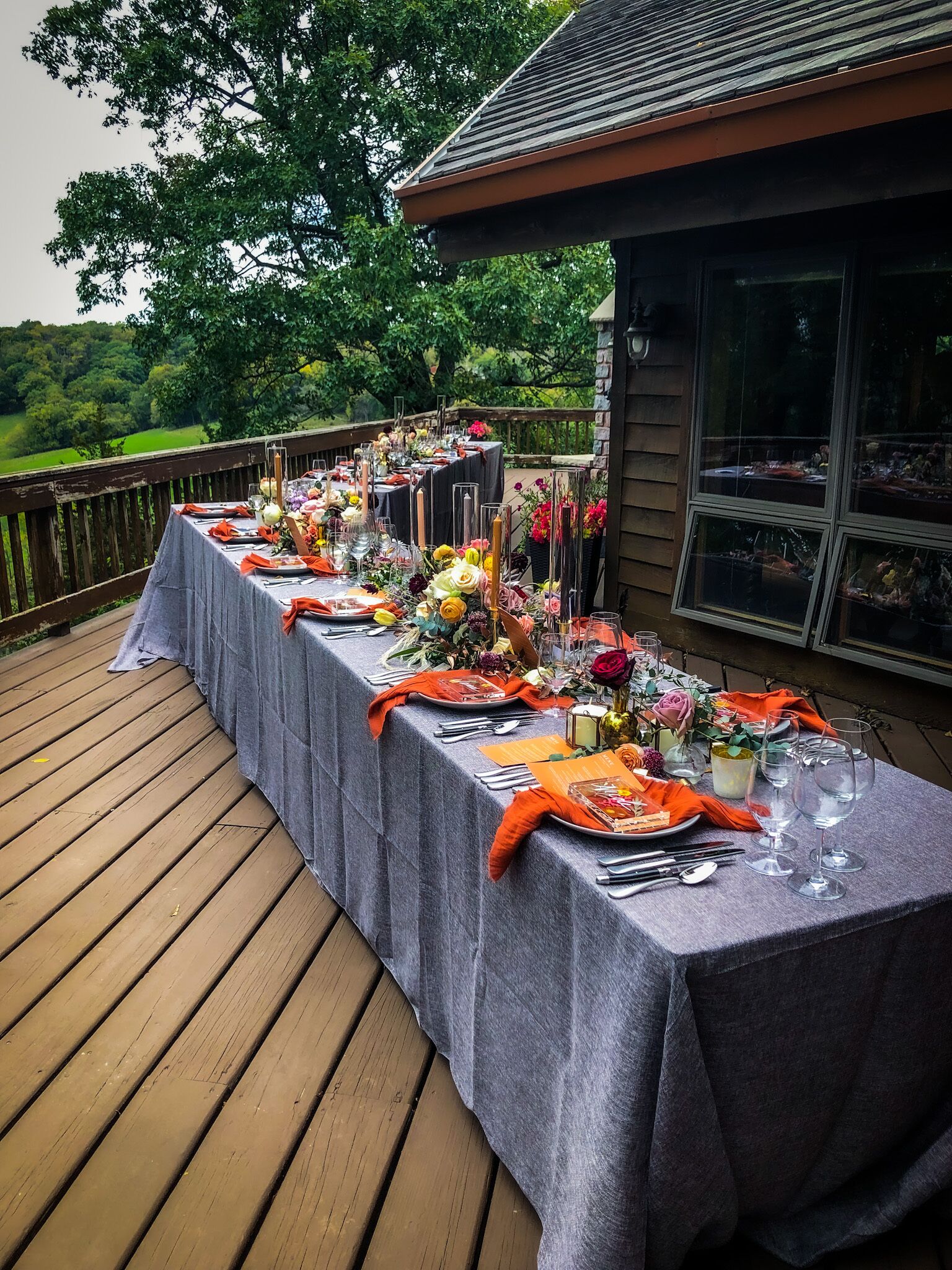 Een buitenterras met een lange tafel gedekt voor een maaltijd, omgeven door een houten terras en een gebouw met grote ramen.