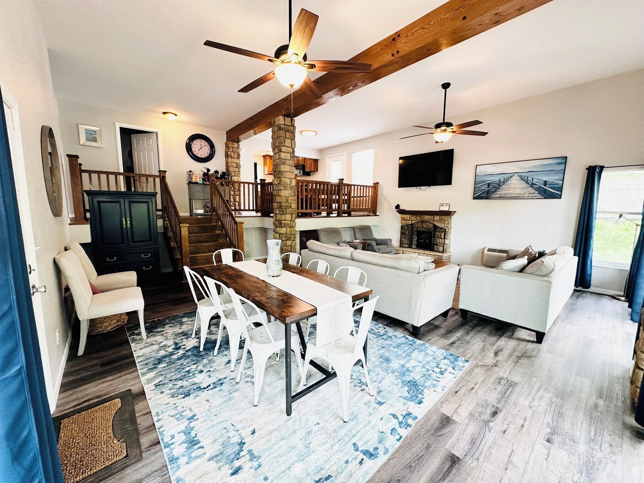 A spacious living area with a dining table, white chairs, a sofa, and a fireplace. There is a staircase leading to an upper level, and a TV mounted on the wall.