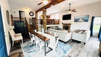 A spacious living area with a dining table, white chairs, a sofa, and a fireplace. There is a staircase leading to an upper level, and a TV mounted on the wall.