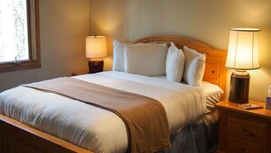 Een net opgemaakt bed met witte lakens en een bruine plaid, met aan beide zijden een bedlampje en een nachtkastje met zakdoekjes.