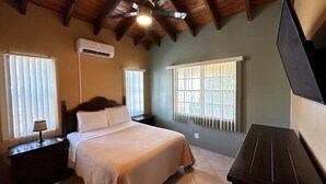 Ein Schlafzimmer mit einem Bett, einem Nachttisch, einer Lampe, einem Fenster mit Gitterstäben, einer Klimaanlage und einem Deckenventilator.