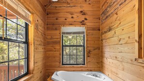 A wooden cabin bathroom with a large bathtub, a window with blinds, and a view of trees outside.