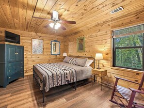 A wooden cabin bedroom with a bed, nightstand, lamp, and a window with a view of trees. - Country Charm B - Pet Friendly Cabin in city limits of Helen GA (Helen)