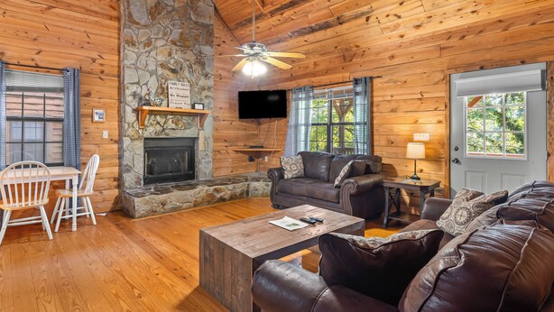A cozy cabin interior with a stone fireplace, wooden walls, a ceiling fan, a TV, and a door with a window.