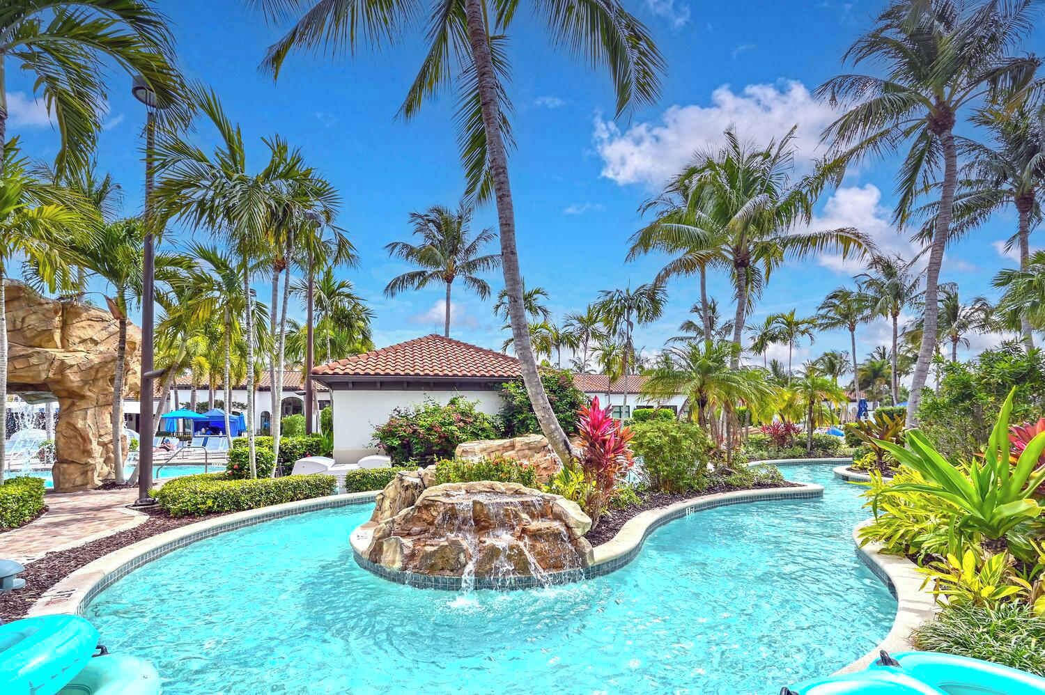 A resort pool with palm trees, a rock waterfall, and a building with a tiled roof.