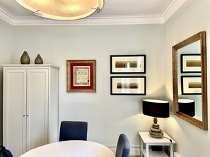 A room with a white cabinet, a framed certificate, two framed pictures, a mirror, and a table with a lamp.