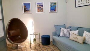 Uma sala de estar com uma poltrona marrom, um banquinho azul, uma mesa clara e um sofá azul com almofadas estampadas.
