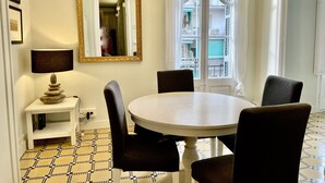Uma área de refeições com uma mesa redonda, quatro cadeiras, um abajur, uma pequena mesa lateral, um espelho e vista para varandas através de uma janela.