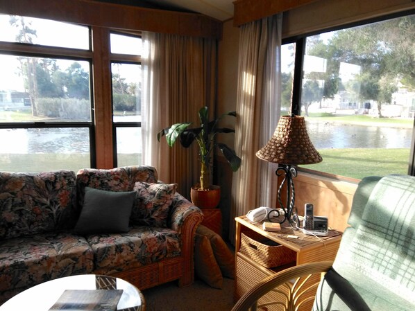 Une pièce avec un canapé à motifs floraux, une petite table, une lampe et une vue sur un lac, visible à travers de grandes fenêtres.