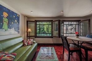 A room with a green sofa, a dining table with chairs, a window with a view of greenery, and a wall with a clock and a painting.