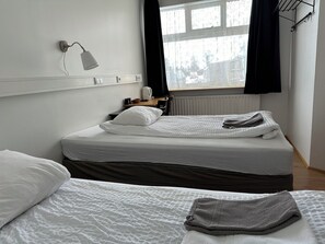 A hotel room with two beds, a window with black curtains, a bedside table with a lamp, and a radiator. - Kanslarinn Hostel (Rangárþing ytra)