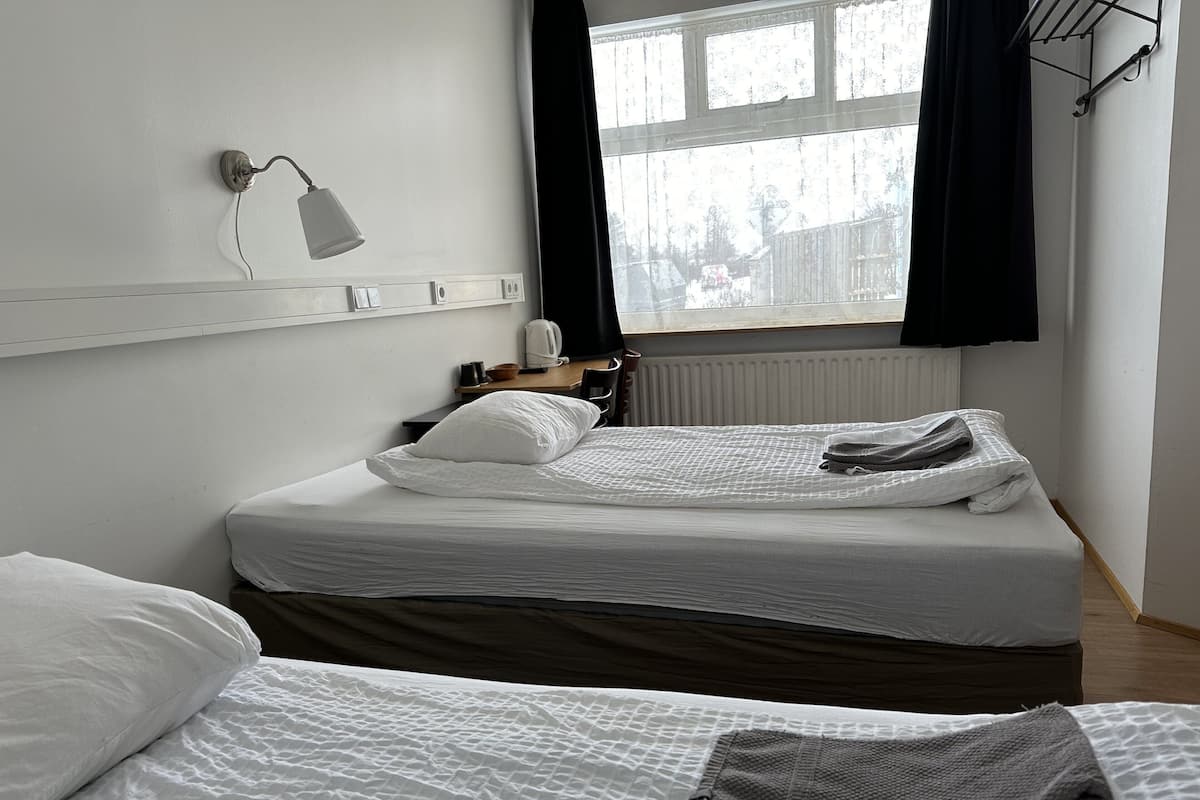 Quarto de hotel com duas camas, uma janela com cortinas pretas, mesa de cabeceira com abajur e radiador.