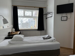 A neatly made bed with white bedding, a bedside table with a lamp, a radiator, a window with black curtains, and a mounted television. - Kanslarinn Hostel (Rangárþing ytra)