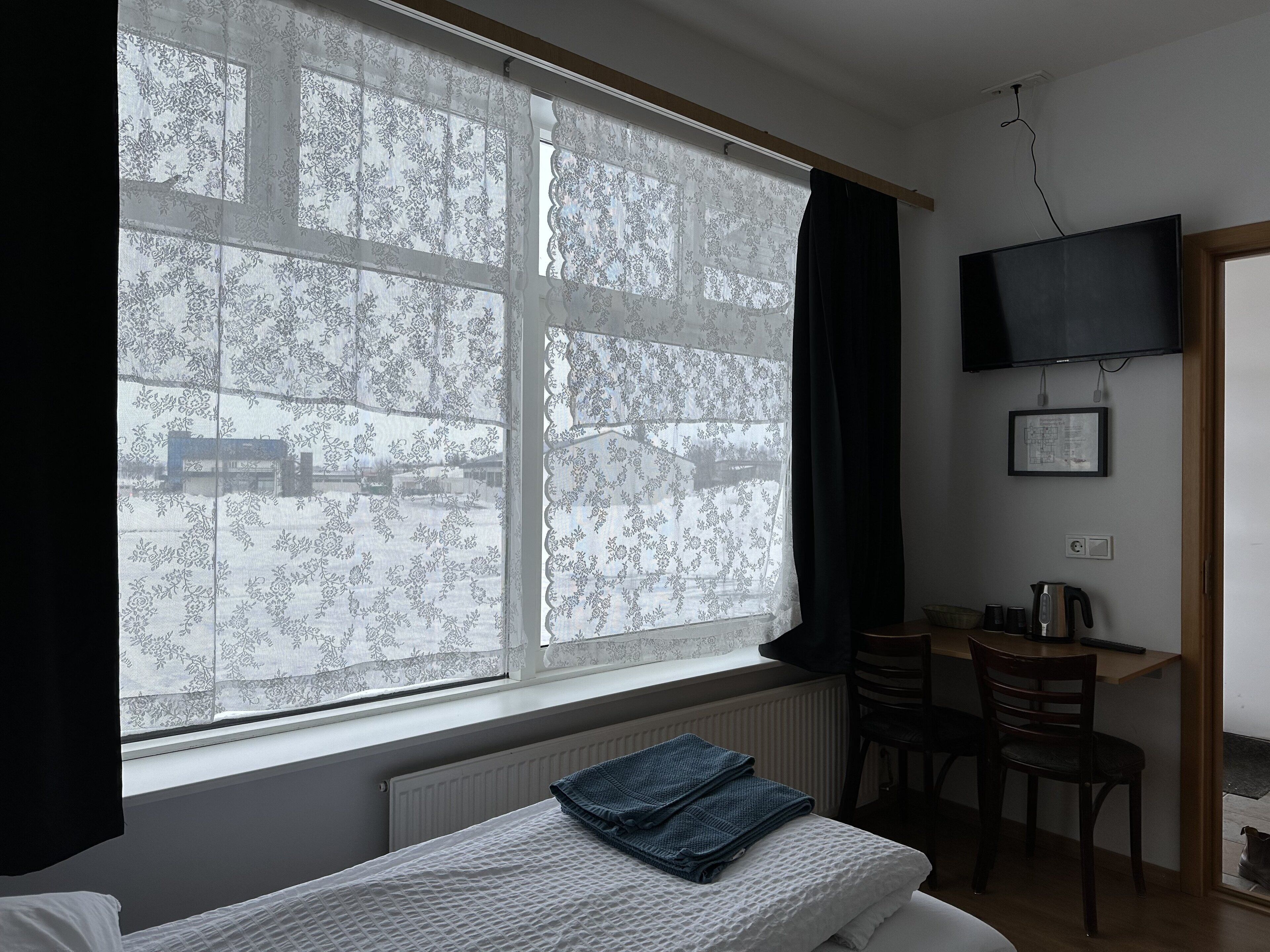 Una habitación con un ventanal grande, una cama, un escritorio con una silla, un televisor montado en la pared y un radiador debajo de la ventana.