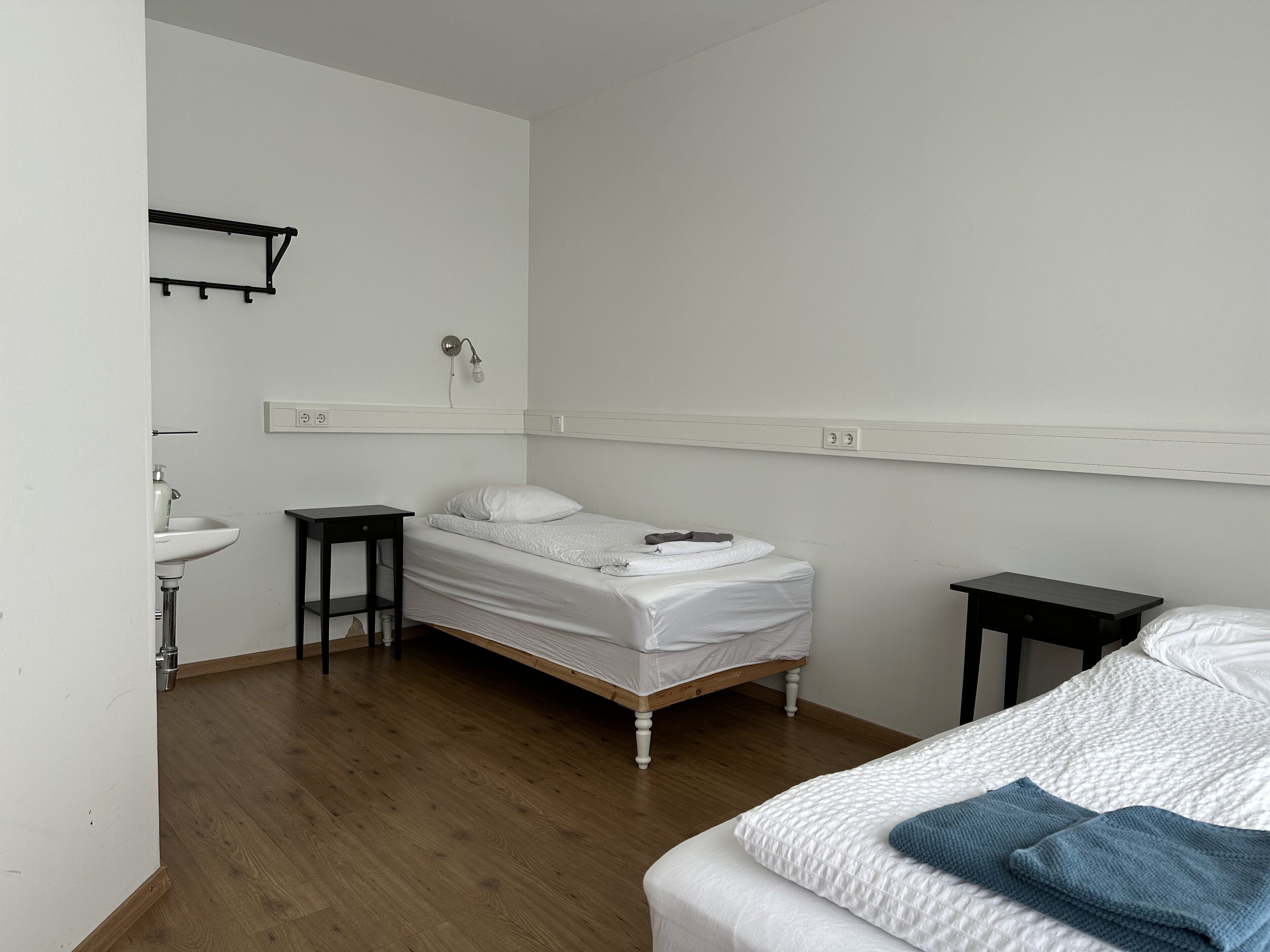 Une petite pièce propre, avec un lit simple, une table de chevet, une commode, une étagère fixée au mur et un lavabo.