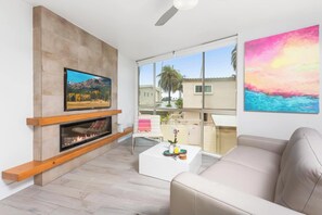 A modern living room with a fireplace, a flat-screen TV, a sofa, and a balcony with a view of palm trees.